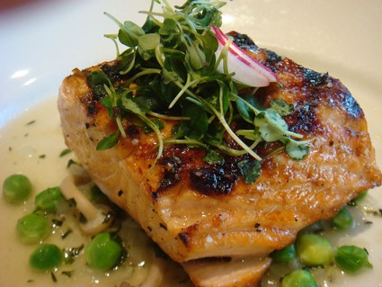Close-up of a grilled salmon fillet topped with microgreens and radish, served with peas and sauce.