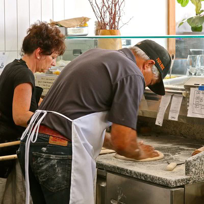 Two people preparing wine grapes at a winery, one kneading grapes on a stone surface, the other observing, in a bright, clean.