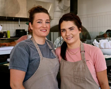 Two women in aprons smiling in a kitchen, representing Lodi Winegrape Commission.