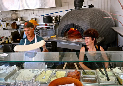 Two people preparing pizza in a commercial kitchen with a large wood-fired oven in Lodi, California.