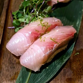 Close-up of two fresh sushi nigiri with fish slices on rice, garnished with green herbs on a wooden surface.