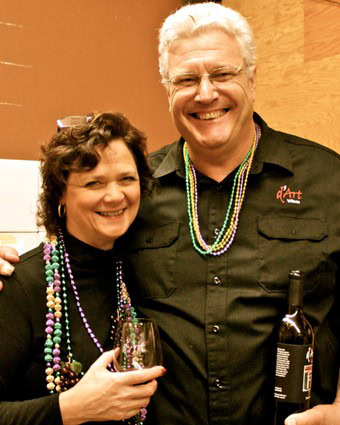 Two smiling individuals at a wine event, wearing colorful beads, holding wine glasses and a bottle.