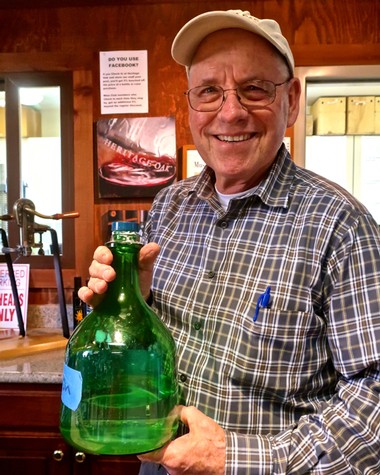Smiling man holding a large green glass bottle in a winery setting, wearing glasses, a cap, and a plaid shirt.