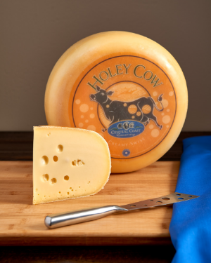 Holey Cow cheese wheel with a slice cut, showcasing its creamy texture and holes, on a wooden surface with a blue cloth.
