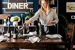 Woman pouring wine at a table with bottles and glasses, in a cozy restaurant setting.