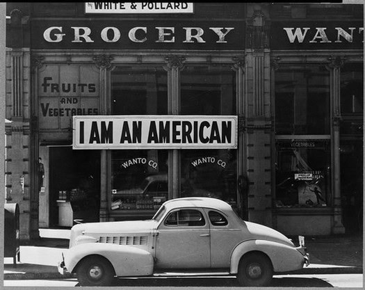 Vintage black and white photo of a grocery store with a classic car parked in front and a sign reading "I AM AN AMERICAN".