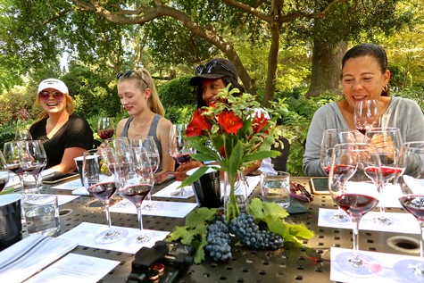 Group of women enjoying wine tasting outdoors with glasses of red wine and a floral centerpiece.