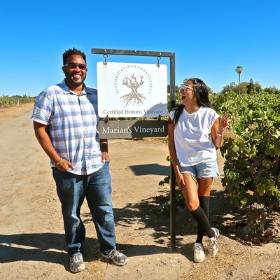 Two people smiling at Marian's Vineyard sign in Lodi, California, with grapevines and clear blue sky.