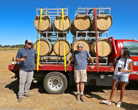 Three people standing beside a red truck loaded with wine barrels during a sunny day in Lodi, California.