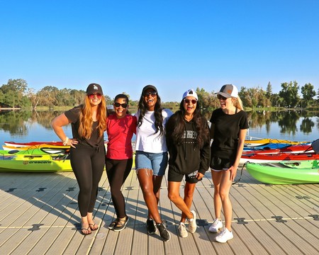 Five women standing on a dock by the lake with colorful kayaks, smiling and enjoying a sunny day outdoors.