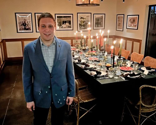 Young man in a blue suit at a dinner event with a candlelit table in the background.