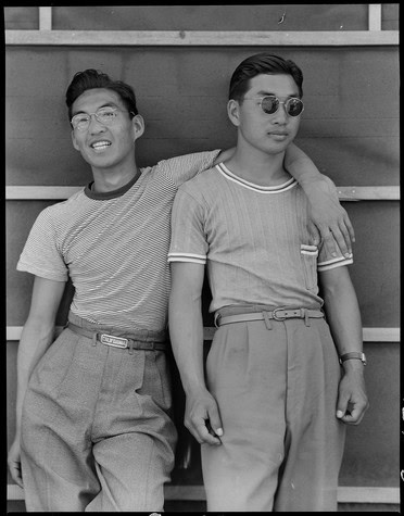 Black and white image of two young men with sunglasses, leaning against a wooden wall, casual style, 1950s.