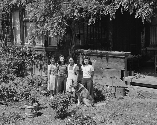 Black and white photo of four women and a dog in a garden with plants and a wooden building in the background.