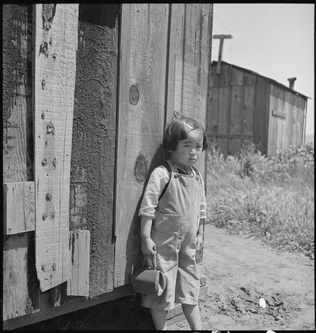 Young girl in overalls holding a hat, standing beside a rustic wooden shed in black and white.
