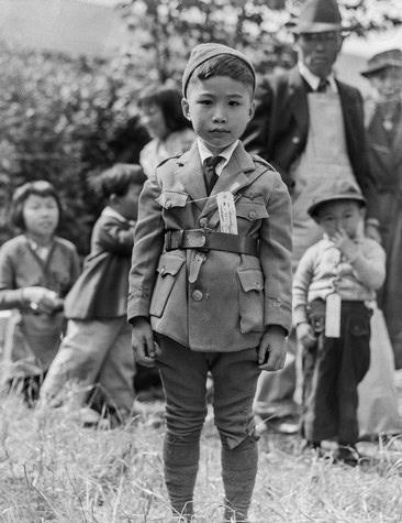 Young boy in vintage military-style coat and cap, standing outdoors with a group of children and adults in the background.
