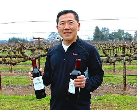 Man holding two bottles of wine in a vineyard, smiling, with grapevines in the background.