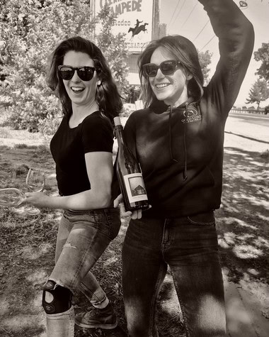 Two women enjoying wine outdoors, holding wine bottles and glasses, smiling and wearing sunglasses.