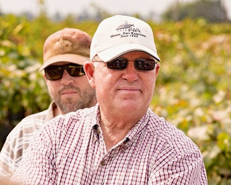 Two men in vineyard, one in foreground wearing sunglasses and white cap, the other in background with hat and sunglasses.