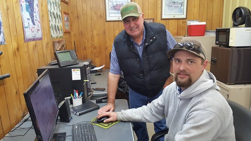 Two men in an office with computers and wood-paneled walls, discussing winegrape industry topics.