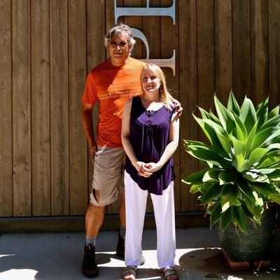 Two people standing outdoors in front of a wooden wall with a large green plant, smiling at the camera.