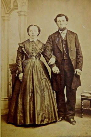 Sepia-toned photo of a man and woman in 19th-century attire, standing indoors with ornate architectural details.