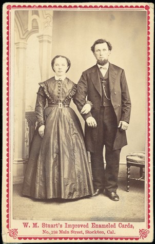 Vintage photo of a man and woman in 19th-century attire, standing indoors with ornate architecture in the background.