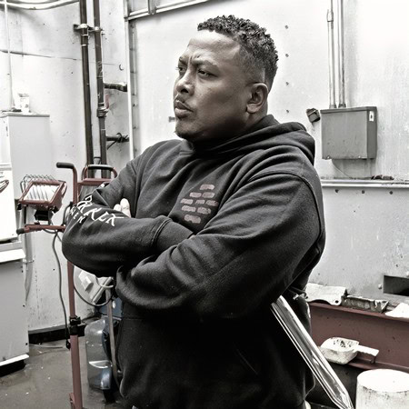Man with crossed arms in a black hoodie, in an industrial environment with tools and equipment.