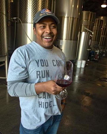 Smiling man holding a glass of red wine in a winery with stainless steel tanks in the background.