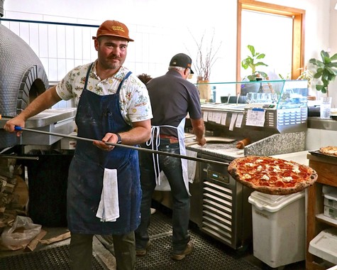 Two men preparing pizza in a kitchen, one holding a pizza peel, with a pizza ready to bake in the foreground.