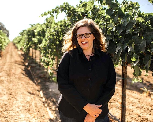 Woman smiling in a vineyard with lush grapevines on sunny day, promoting Lodi Winegrape Commission.