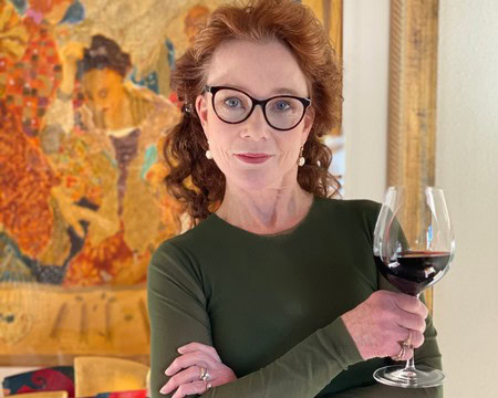 Woman with glasses holding a glass of red wine in a winery setting, with colorful artwork in the background.