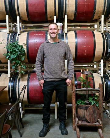 Smiling man in winery with stacked wine barrels and grapevines, promoting Lodi wine tourism and winegrape industry.