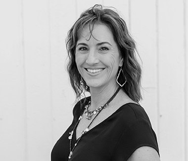 Smiling woman with wavy hair wearing a black top and jewelry, portrait in black and white.