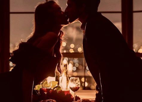 Silhouette of a couple kissing at sunset with wine glasses on table, romantic scene, warm lighting, city lights in background.
