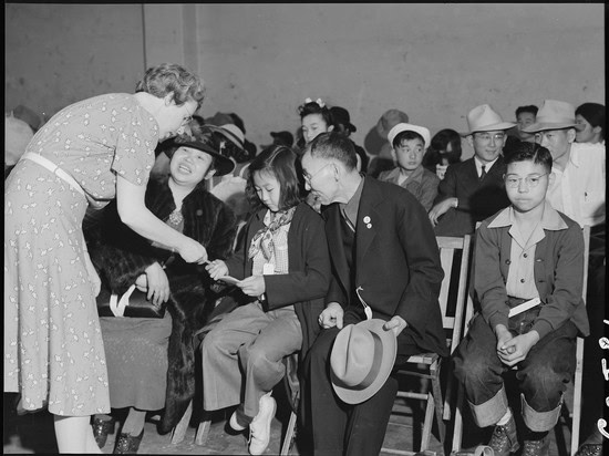 Historical black and white photo of people at a formal event, with a woman handing an award to a seated man.
