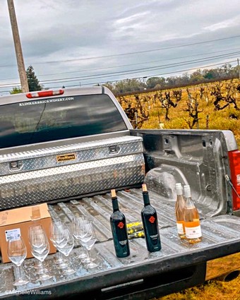 Wine bottles and glasses in truck bed at Lodi vineyard with grapevines in background.
