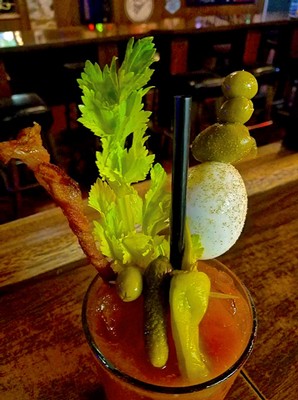 Creative cocktail with green garnishes, bacon strip, and skewered olives on a wooden bar counter.