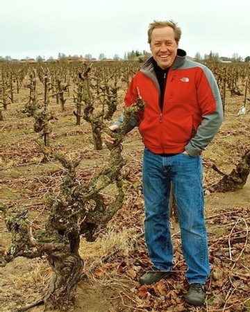 Smiling man in red jacket standing in a vineyard with pruned grapevines in Lodi, California.