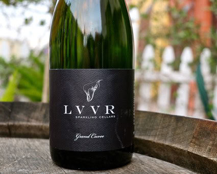 Close-up of a LUVR Sparkling Cellars wine bottle on a wooden surface with a garden background.