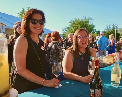 Two women enjoying wine at a Lodi Winegrape Commission outdoor event with a crowd in the background.