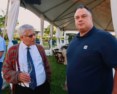 Two men at a Lodi Winegrape Commission event, one holding a wine glass, under a large outdoor tent.