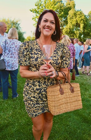 Woman smiling at outdoor wine tasting event holding a glass of wine and a wicker handbag.