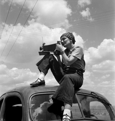 Black and white photo of girl sitting on car roof with camera, under cloudy sky, vintage style.