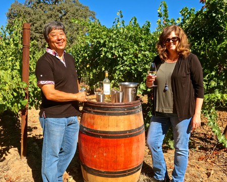 Two women enjoying wine tasting outdoors at Lodi Winegrape Commission vineyard with a wine barrel and grapes in background.