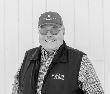Smiling man in Lodi Winegrape Commission cap and vest, standing outdoors.