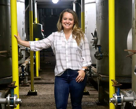 Woman smiling in a winery with industrial tanks and yellow pipes, representing Lodi Winegrape Commission.