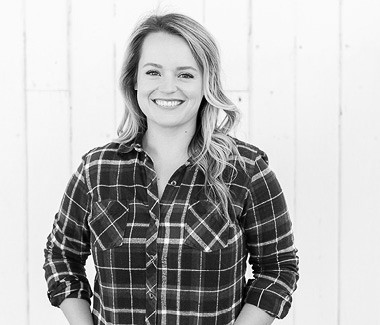 Smiling woman in plaid shirt standing outdoors with a wooden fence background.