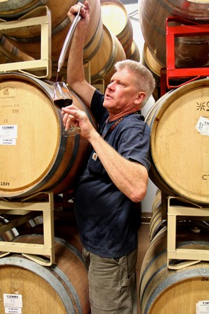 Man tasting wine from a barrel at a winery, surrounded by wine barrels in a cellar.