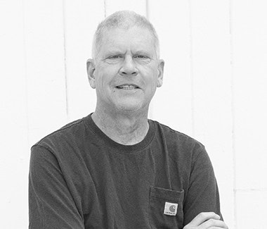 Black and white portrait of a middle-aged man with short hair, wearing a dark t-shirt, standing outdoors.