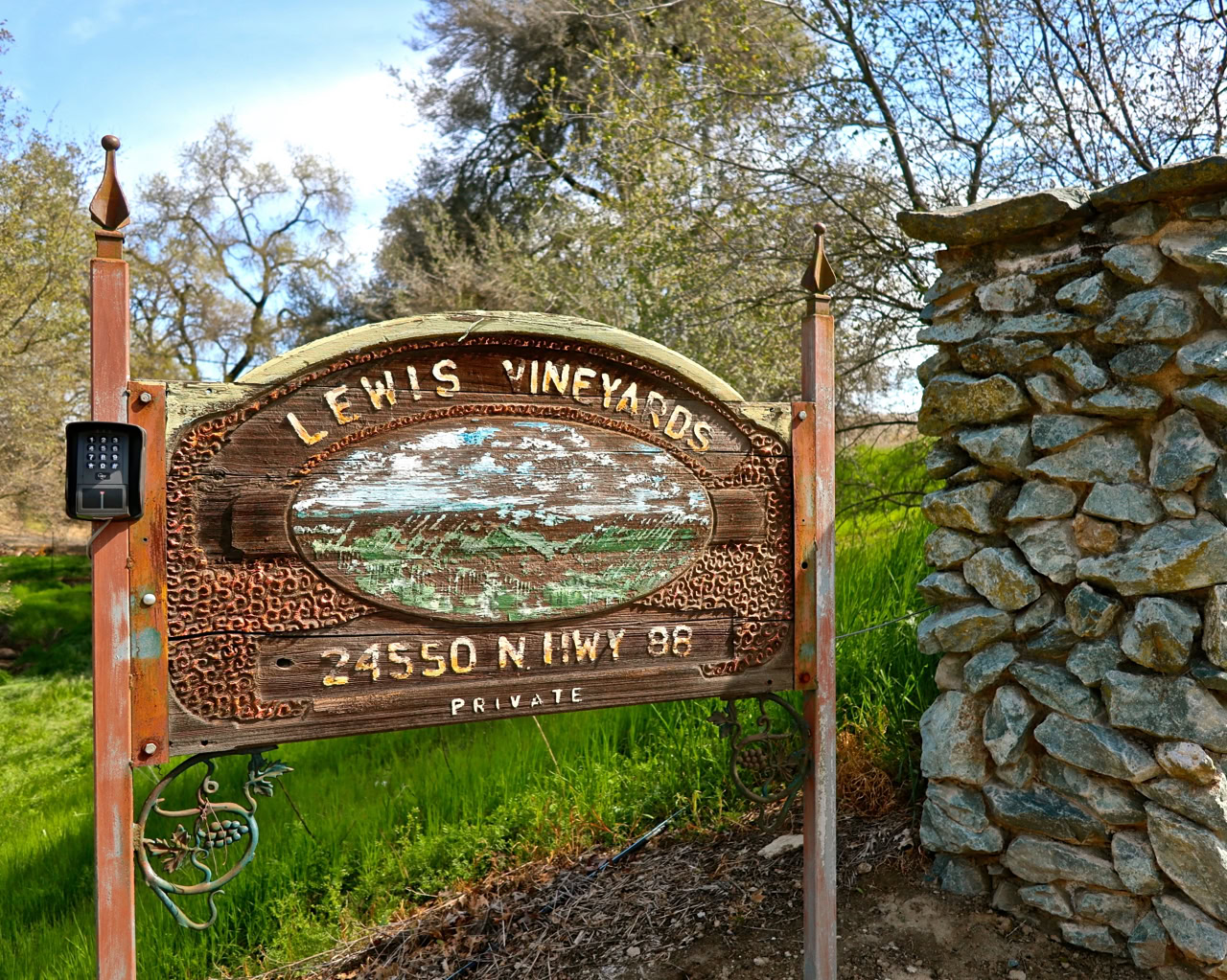 Rustic wooden sign for Lewis Vineyards at 24550 N Hwy 99 in Lodi, California, surrounded by greenery and stone wall.